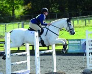 Pictured is a grey horse and rider showjumping over an over. The horse is wearing protective boots on their front legs