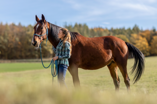 Pictured is a female rider standing next to a bay horse in a field; natural horsemanship can do wonders for your bond